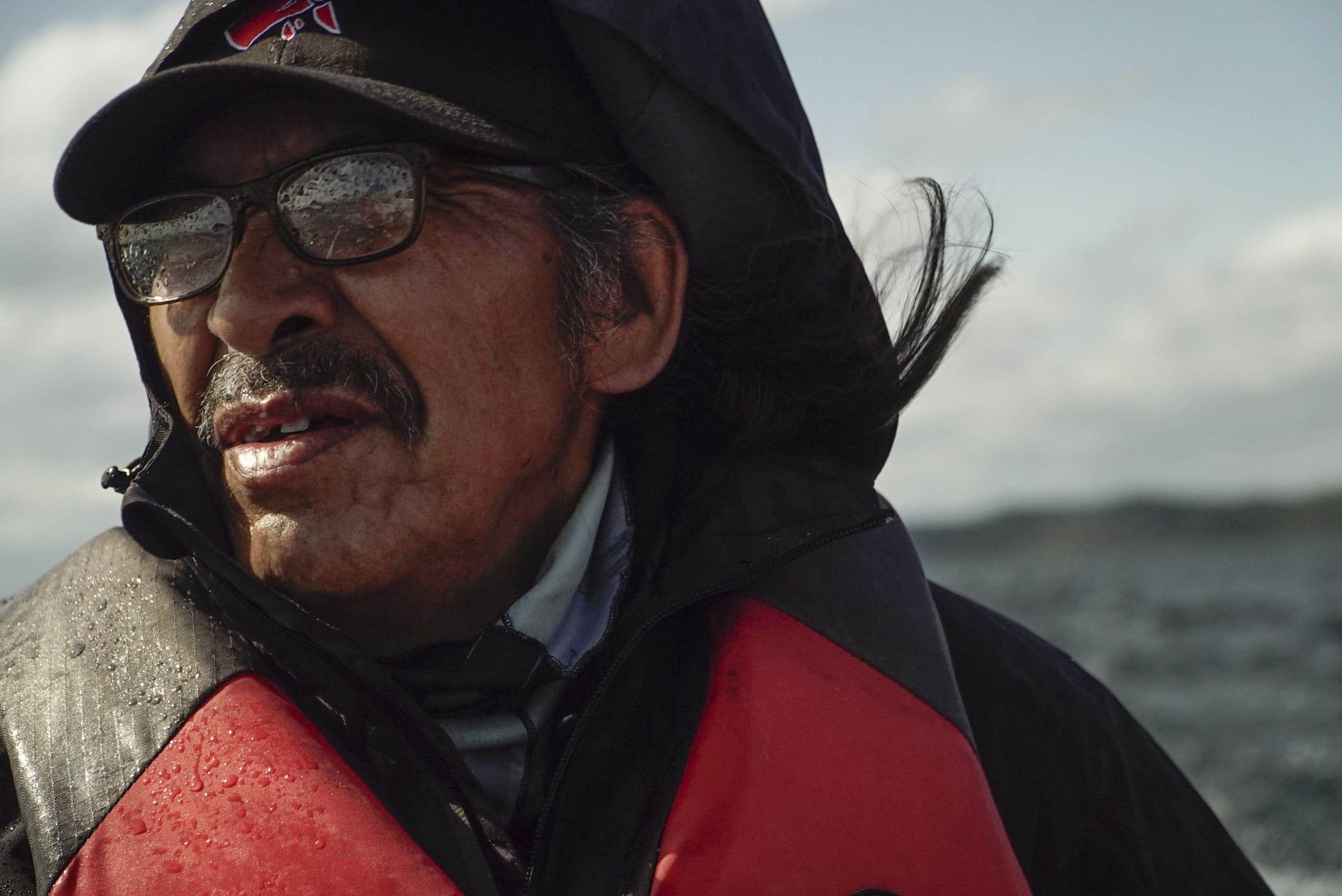 A portrait of Łutsël K’é Dene community member and Thaidene Nëné management board member, JC Catholique. JC appears to be on a boat and is wearing sunglasses, an orange life jacket and black ball cap. There is water in the background and blue skies.
