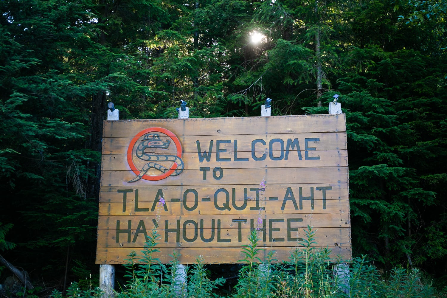 Photo of a wooden sign that reads “Welcome to Tla-o-qui-aht Ha’houlthee”. The sign is in front of green cedar trees.