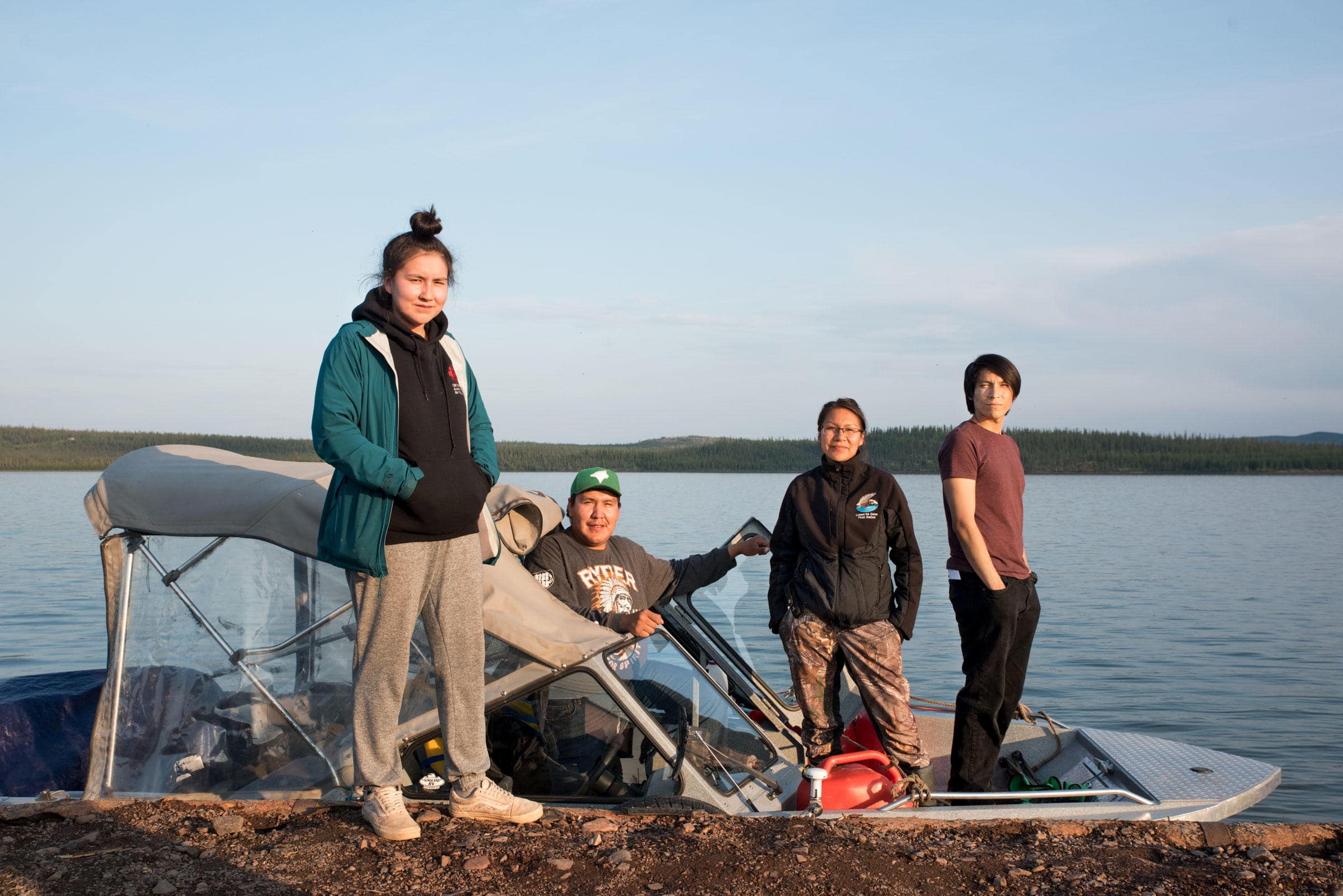 Three Łutsël K’é Dene Youth Guardians are on a boat with water in the background and land on the horizon. One Youth Guardian is standing in front of the boat on a large rock.
