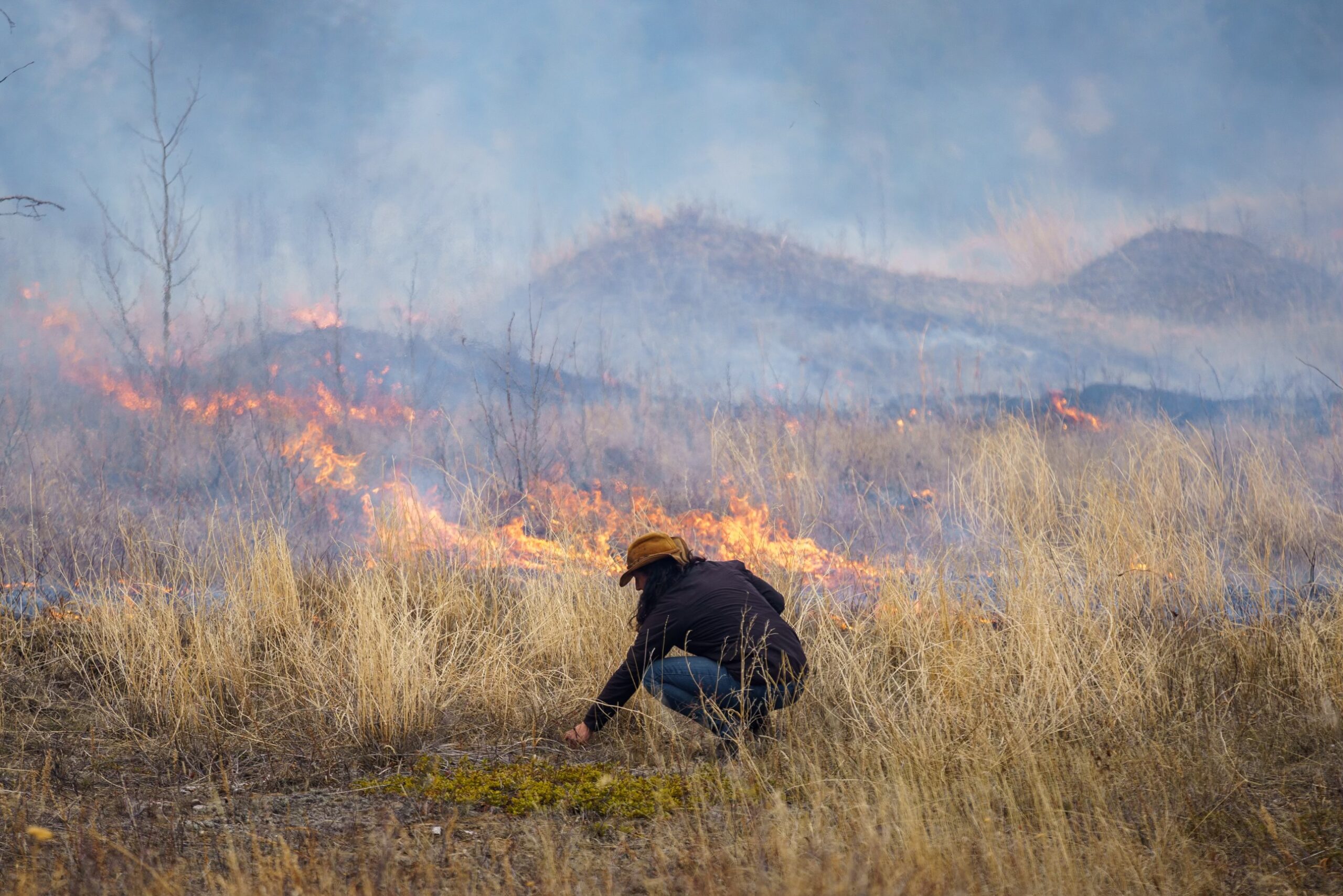 A person with their back to the camera crouches in a field near a perscribed, traditional fire.